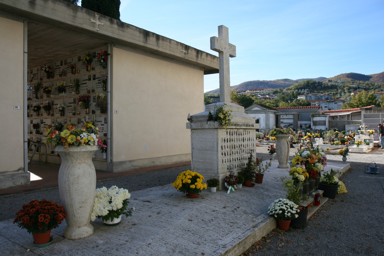 Monumento funerario con fiori e croce, circondato da un cimitero e colline sullo sfondo.