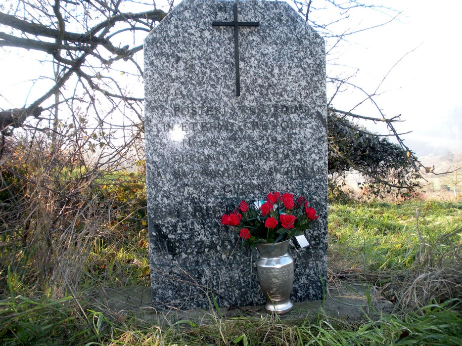 Cippo commemorativo con croce, fiori e iscrizioni, circondato dalla natura.