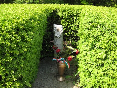 Cippo commemorativo circondato da siepi verdi, con fiori e una decorazione tricolore.