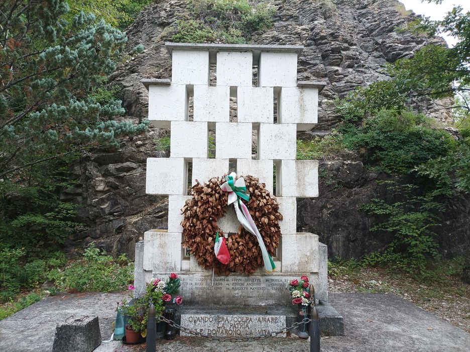 Monumento ai caduti di Rio Farnese, con una corona di fiori e decorazioni, circondato da natura.