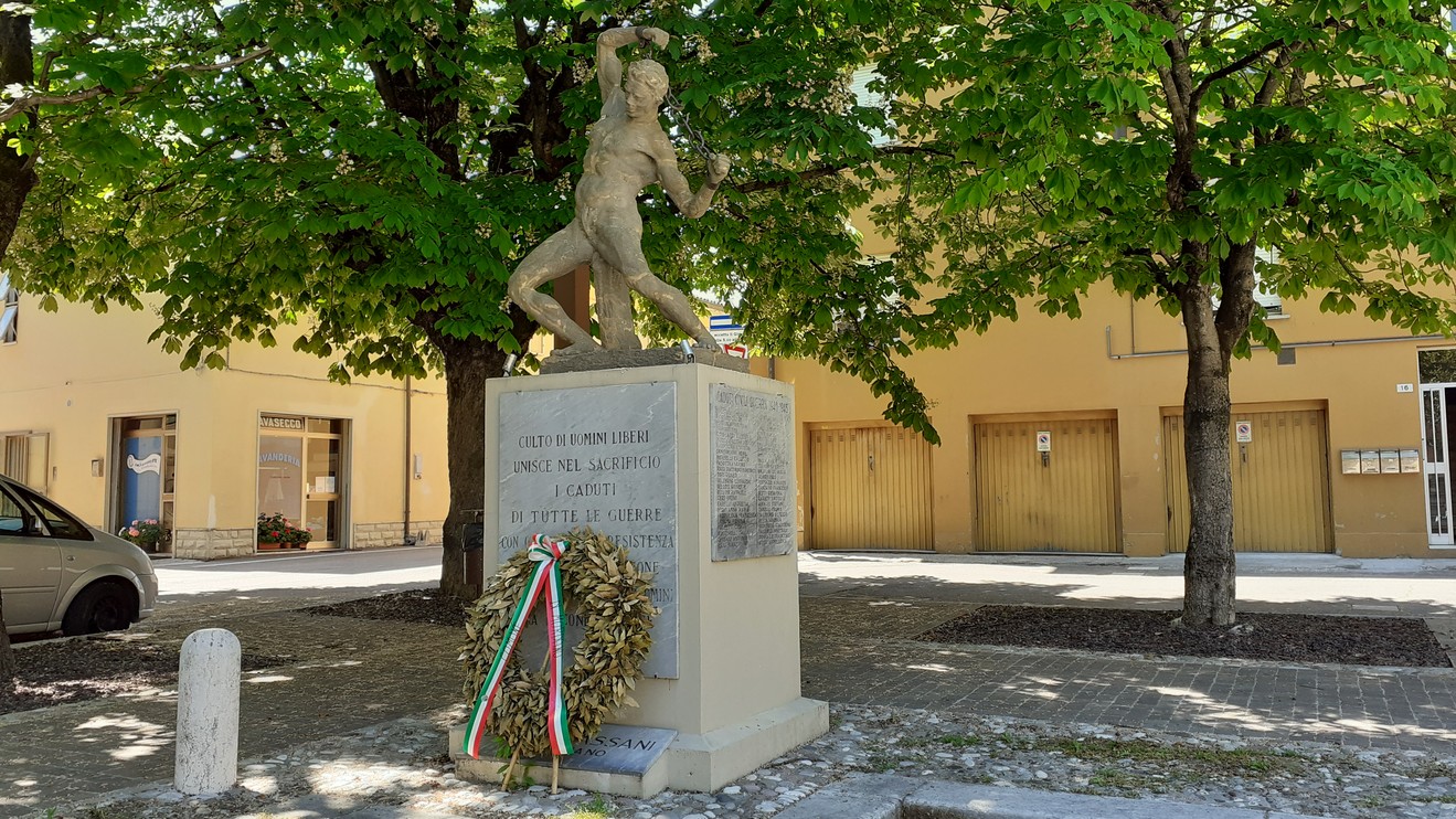 Monumento ai caduti di Mordano, con statua centrale e corona di fiori, circondato da alberi verdi.