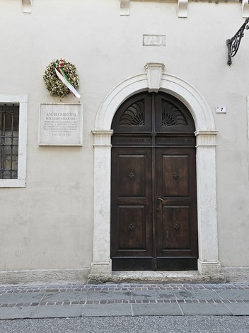 Lapide commemorativa su una parete accanto a una porta in legno, decorata con una corona di fiori.