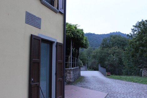 Lapide commemorativa in zona panoramica, circondata da alberi e sentieri che conducono a colline.