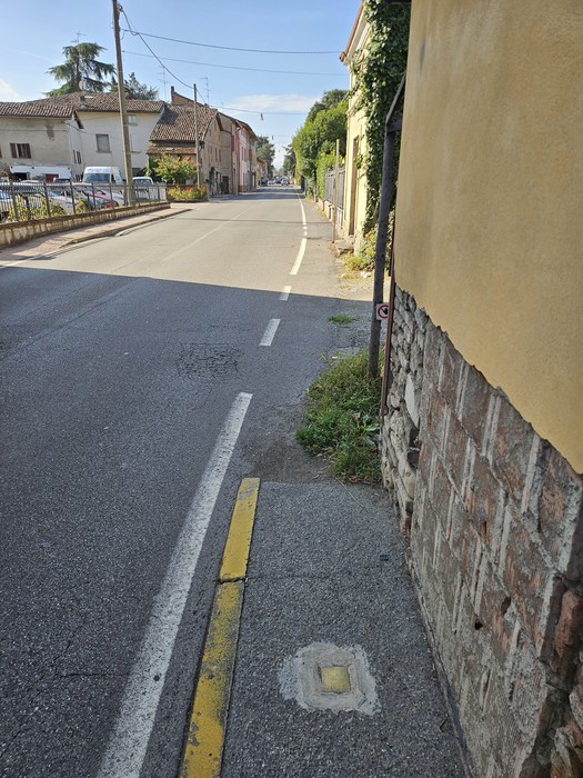 Marciapiede su una strada di periferia, con una piccola pietra d'inciampo incassata nella pavimentazione.