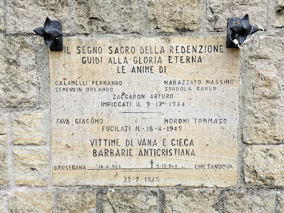 Lapide in pietra con iscrizione, fissata a un muro, sormontata da due decorazioni in metallo scuro.