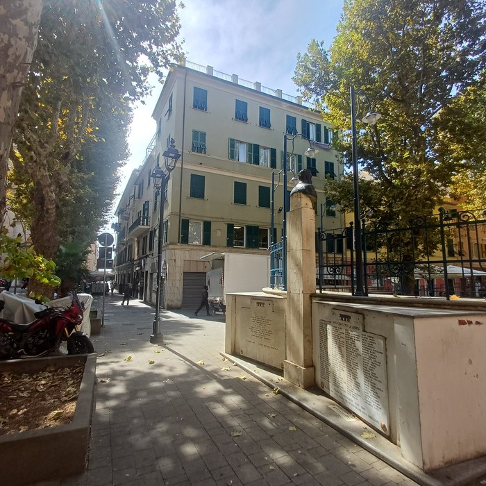 Lapide in pietra con iscrizione su basamento lungo una via alberata, tra edifici cittadini.