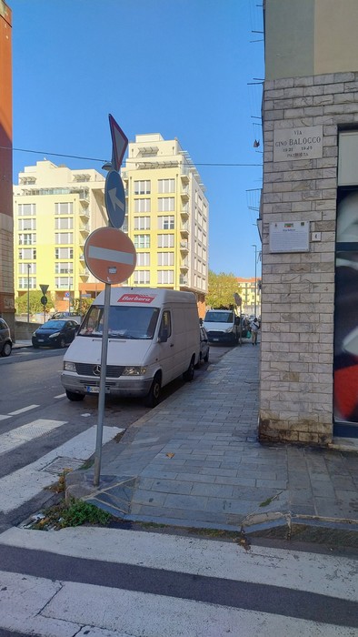 Angolo di strada con edificio in mattoni, targa con iscrizione fissata sulla parete in alto.
