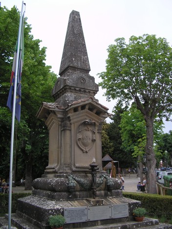 Monumento ai caduti, un obelisco commemorativo in un parco, circondato da alberi e bandiere.