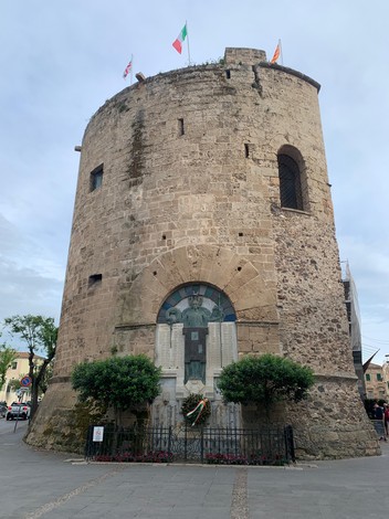 Monumento ai Caduti di Alghero, torre in pietra con decorazioni e bandiere, circondato da verde e un recinto.