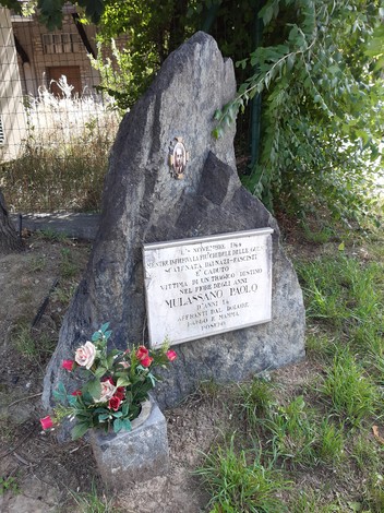 Cippo commemorativo a Mulassano, con decorazioni floreali e incisioni su pietra, circondato da vegetazione.