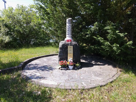 Cippo in pietra con iscrizione su base di roccia, fiori ai piedi, area pavimentata circolare tra alberi.