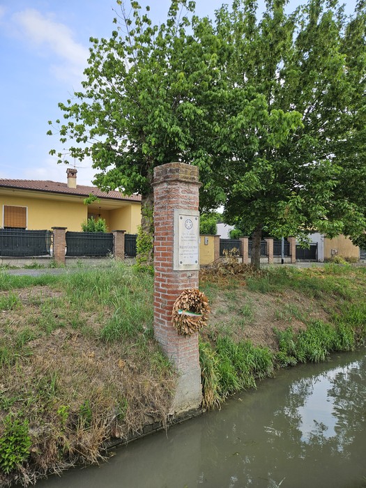 Cippo commemorativo a Cavagnari, immerso nel verde, affiancato da una vegetazione rigogliosa e un canale adiacente.