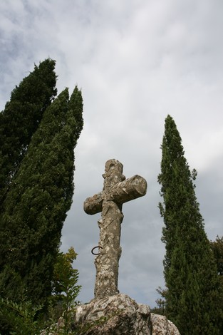 Monumento di Falzano: una croce antica incorniciata da alti cipressi sotto un cielo nuvoloso.