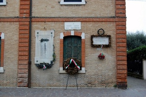 Lapide commemorativa a Gallo, con corone di fiori e decorazioni, su una parete in mattoni.