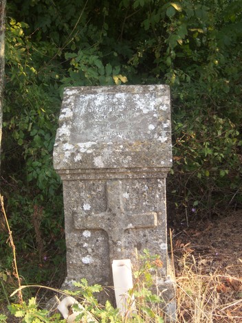 Cippo memoriale in pietra, circondato da vegetazione, con una croce e un design semplice.