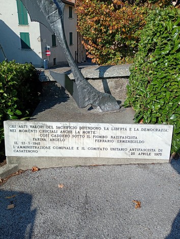 Lapide in marmo con iscrizione, posta su basamento in pietra ai piedi di una scultura, in un giardino.