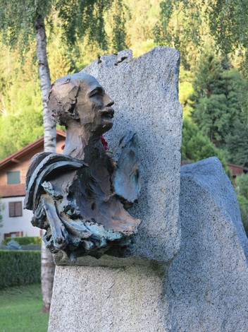 Monumento a Collini, scultura in bronzo su base di pietra, circondata da natura e edifici tipici.