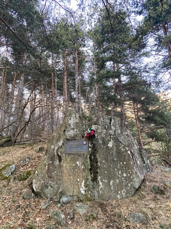 Targa commemorativa su una grande roccia circondata da alberi e vegetazione in un ambiente boschivo.