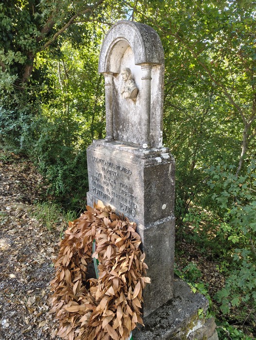 Cippo in pietra con nicchia e rilievo sacro, iscrizione sul basamento, corona secca appoggiata davanti, tra la vegetazione.