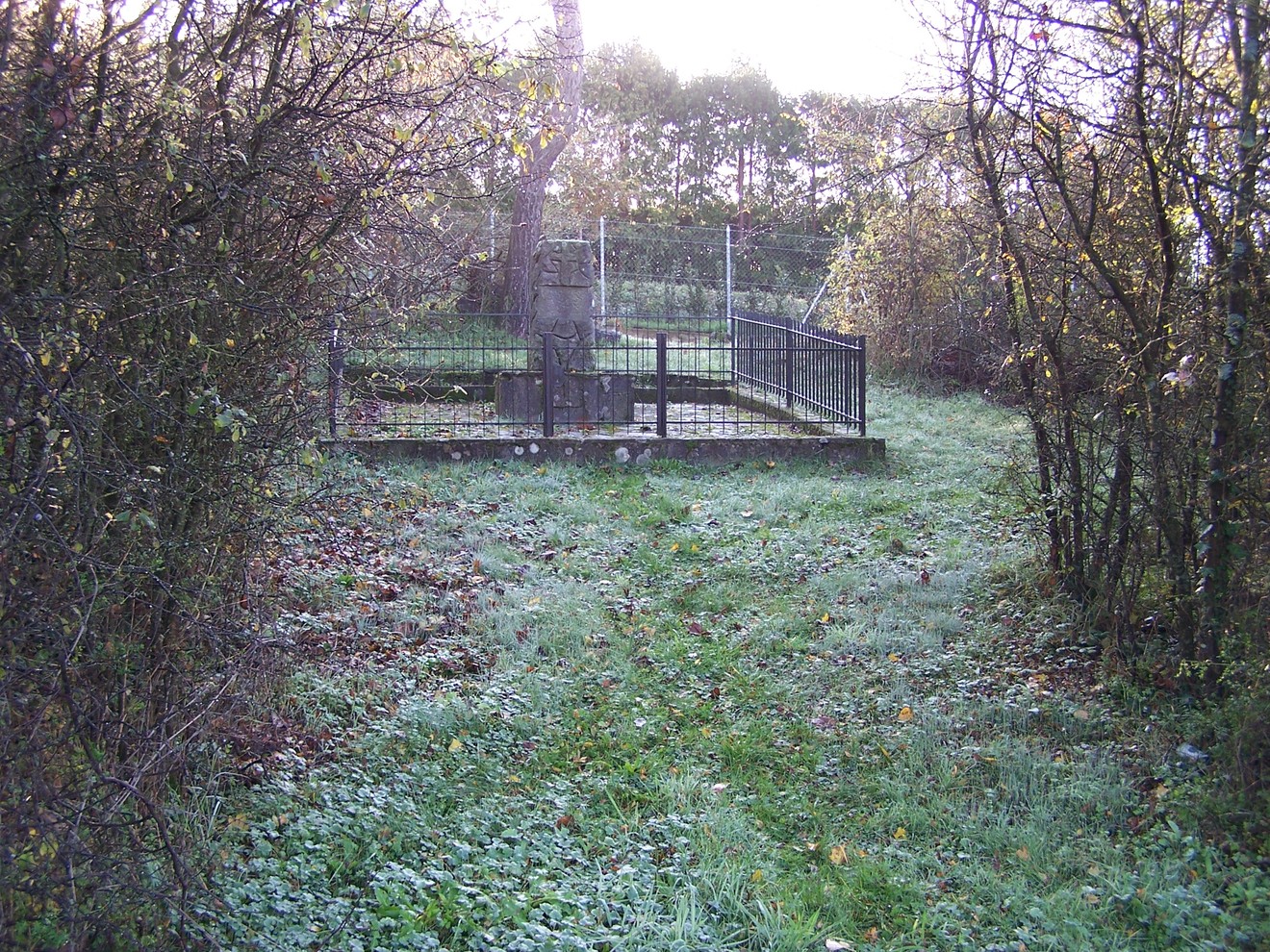 Cippo commemorativo circondato da vegetazione e recinzione, immerso nella tranquillità della natura.
