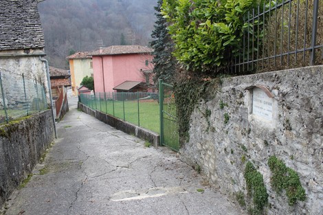 Vista di un viale che conduce a una lapide, circondato da edifici e vegetazione, in un contesto rurale.