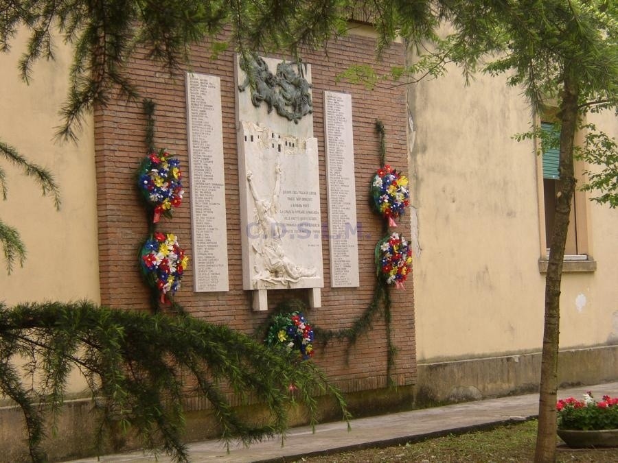 Lapide per le vittime dei bombardamenti, adornata con fiori commemorativi, con un muro di mattoni sullo sfondo.
