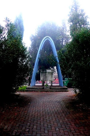 Monumento ai ferrovieri caduti, con arco blu circondato da alberi e percorso lastricato. Atmosfera nebbiosa.
