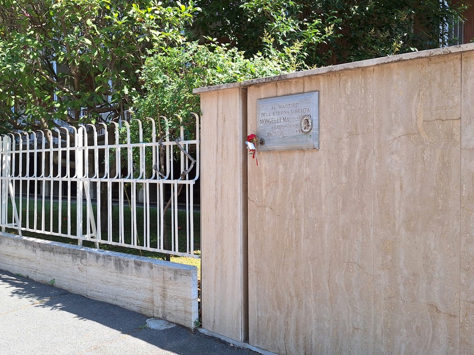 Lapide commemorativa su un muro, con un piccolo omaggio floreale e un cancello sullo sfondo.