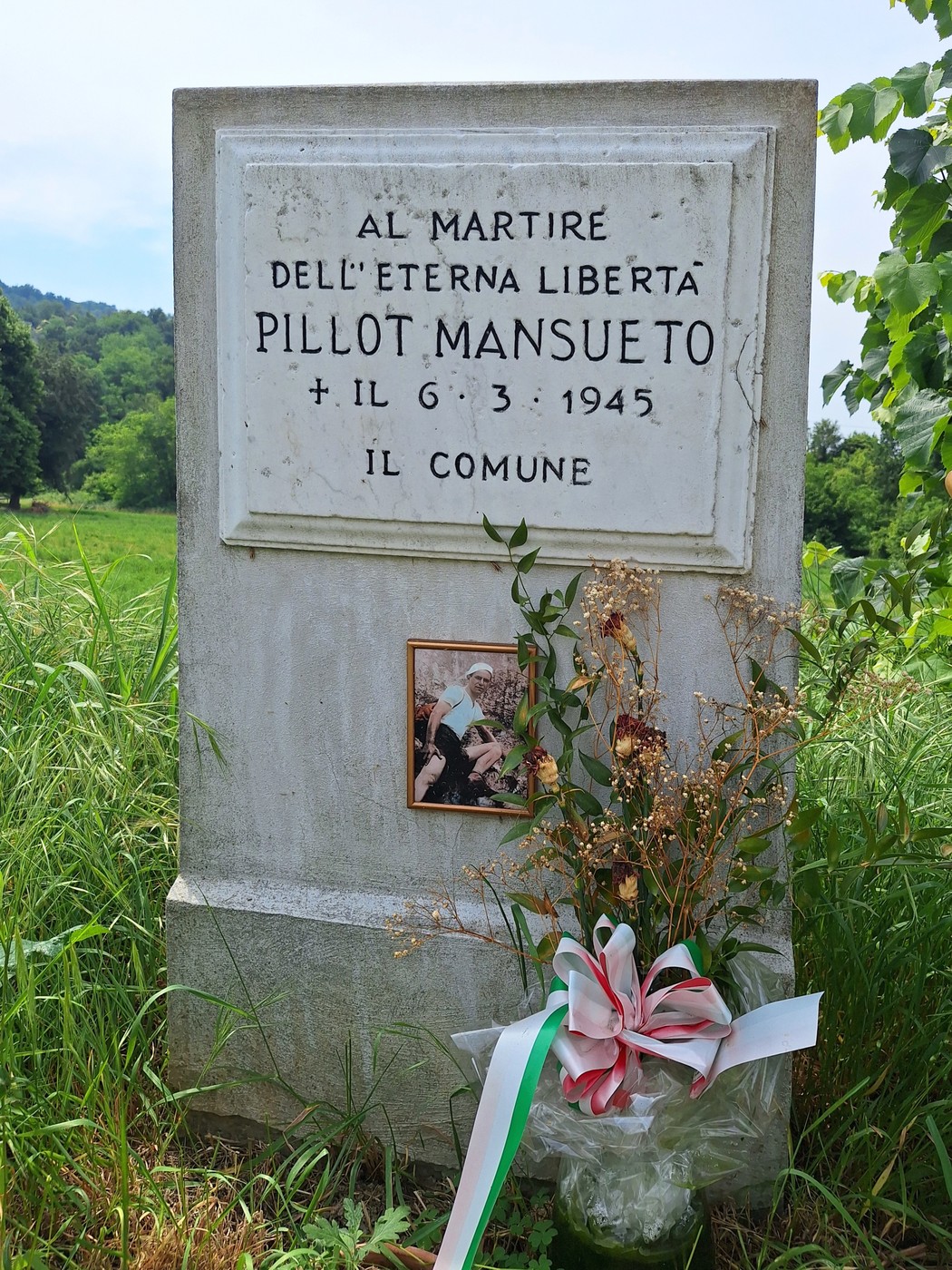 Cippo commemorativo dedicato a Pillot Mansueto, con decorazioni floreali e un'immagine ricordo.