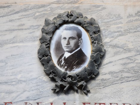 Lapide commemorativa con ritratto di Duò, incorniciato da un festone di edera su sfondo marmoreo.