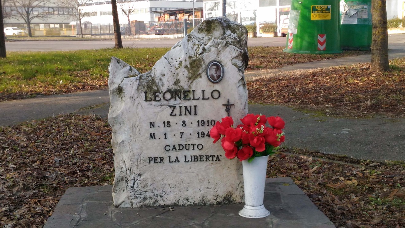 Cippo commemorativo dedicato a Leonello Zini, con fiori rossi, situato in un'area pubblica.