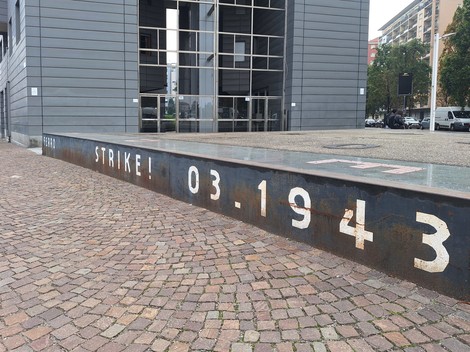 Cordolo metallico con iscrizione incisa, lungo una piazza pavimentata davanti a un edificio a vetrate.