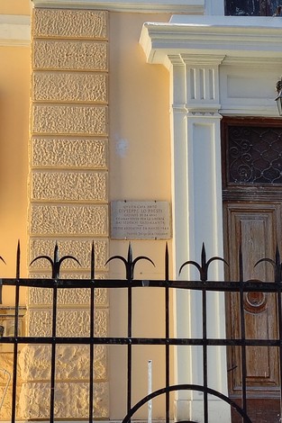 Lapide commemorativa su una parete, vicino a un cancello in un edificio di colore giallo chiaro.