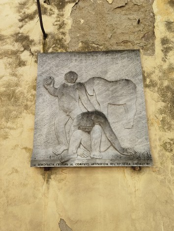 Bassorilievo artistico che rappresenta figure umane, con un forte contrasto su sfondo di parete ingrigita.