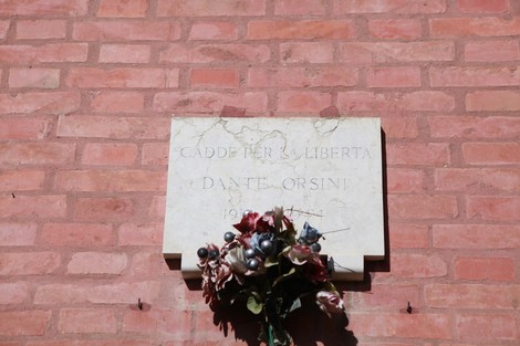 Lapide commemorativa dedicata a Dante Orsini, con decorazioni floreali, su muro di mattoni.