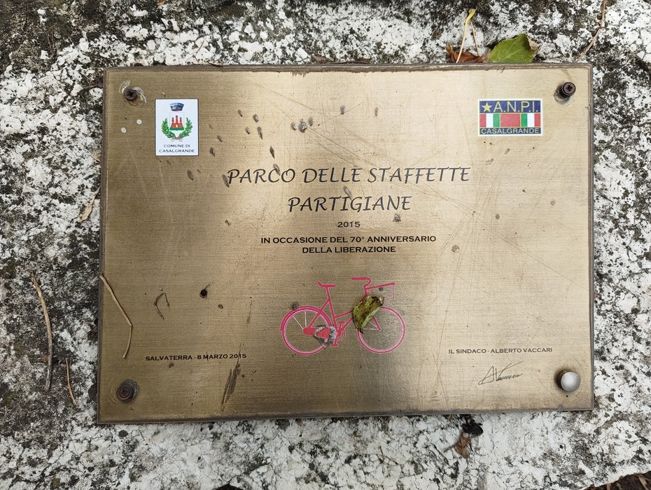 Targa in metallo con iscrizione e disegno di una bicicletta, appoggiata su un masso di pietra.