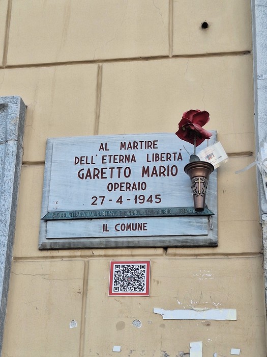 Lapide in marmo fissata a una parete, con iscrizione e un fiore rosso appoggiato accanto.