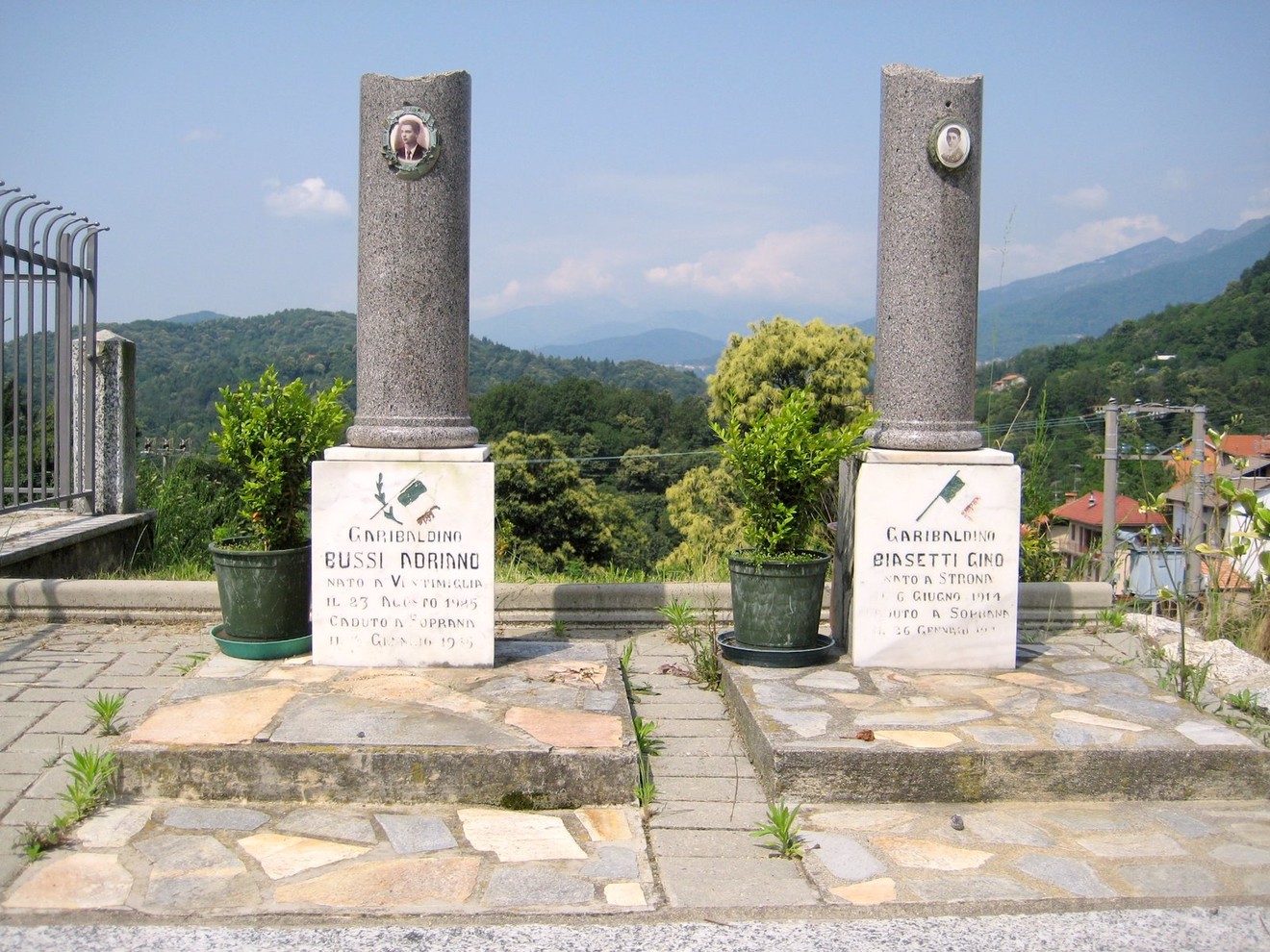 Cippi commemorativi dedicati a Bussi e Biasetti, con sfondo montano e vegetazione lussureggiante.