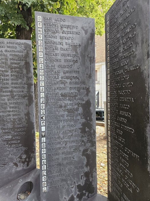 Monumento in lastre di pietra scura, con iscrizioni e piccoli ritratti, disposto all’aperto tra gli alberi.