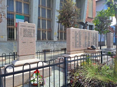 Monumento ai caduti, con iscrizioni commemorative e decorazioni floreali, circondato da una recinzione.