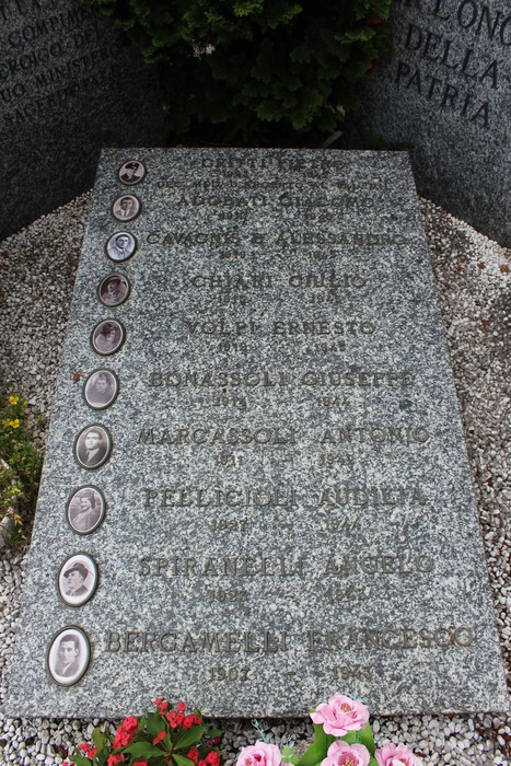 Targa commemorativa con nomi e fotografie dei caduti per la patria, circondata da fiori e piante.