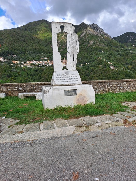 Monumento in pietra bianca con sagoma di un partigiano, basamento con iscrizione, su sfondo collinare.