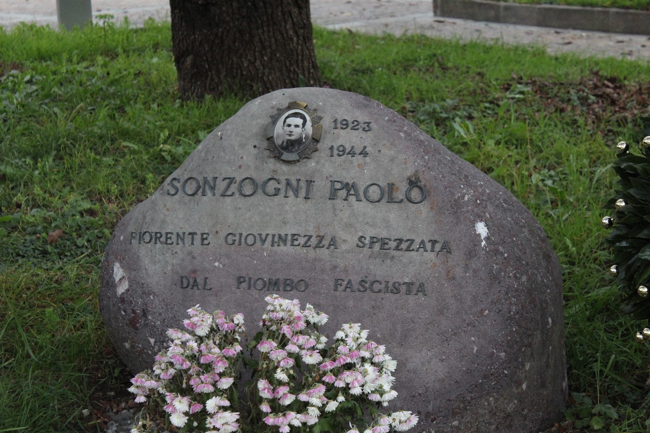 Cippo commemorativo dedicato a Paolo Sonzogni, con foto e data, circondato da fiori.
