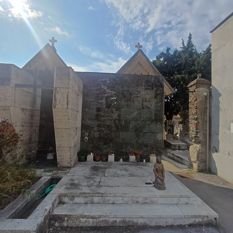 Cappella cimiteriale in pietra con croci sui timpani, vasi di fiori e statua davanti all'ingresso.