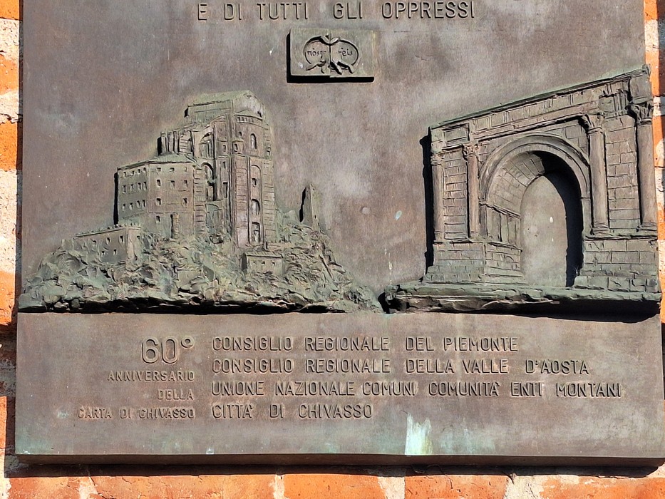 Targa in bronzo su parete con rilievi di un castello e un arco trionfale, sotto un'iscrizione commemorativa.