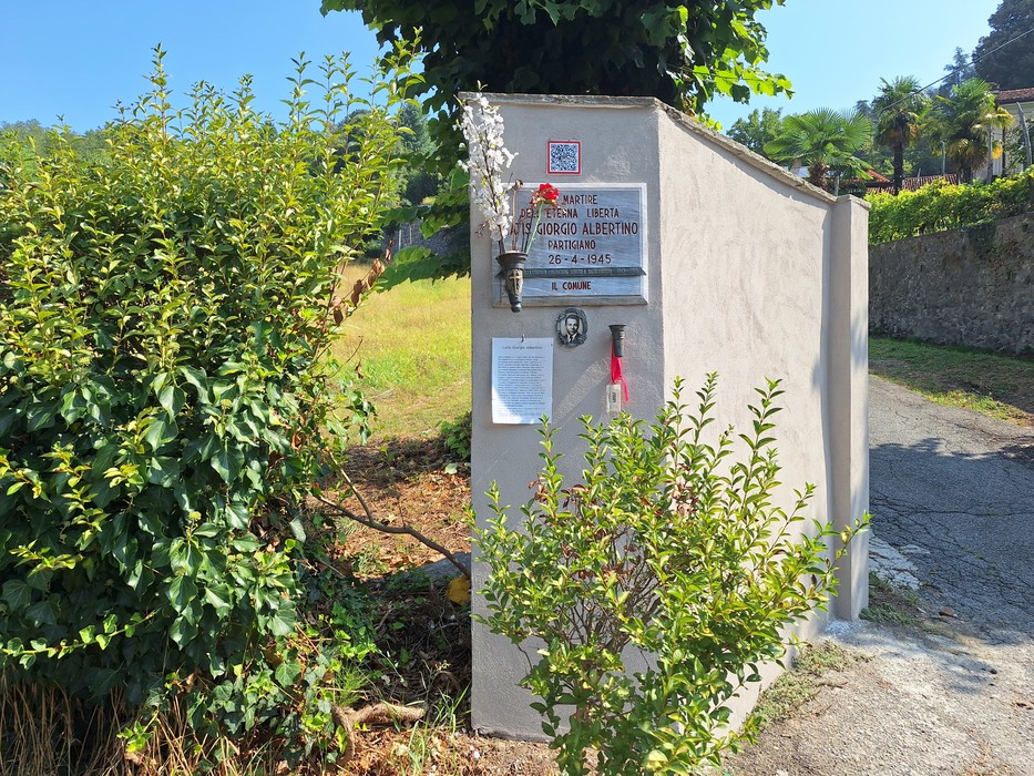 Lapide con iscrizione su parete intonacata ai margini di una strada, circondata da vegetazione.