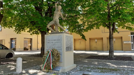 Monumento ai caduti di Mordano, con statua centrale e corona di fiori, circondato da alberi verdi.