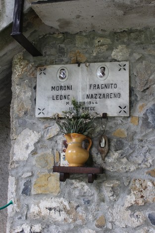 Lapide commemorativa in pietra con nomi e decorazioni floreali, situata su un muro esterno.