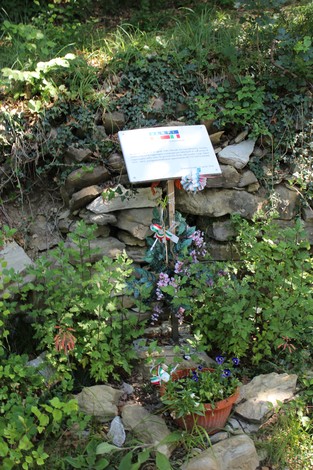Targa commemorativa circondata da piante e fiori, posizionata su una parete di pietra in un contesto naturale.