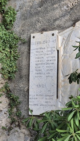 Monumento ai caduti con iscrizioni commemorative su una parete rocciosa, circondato da vegetazione.